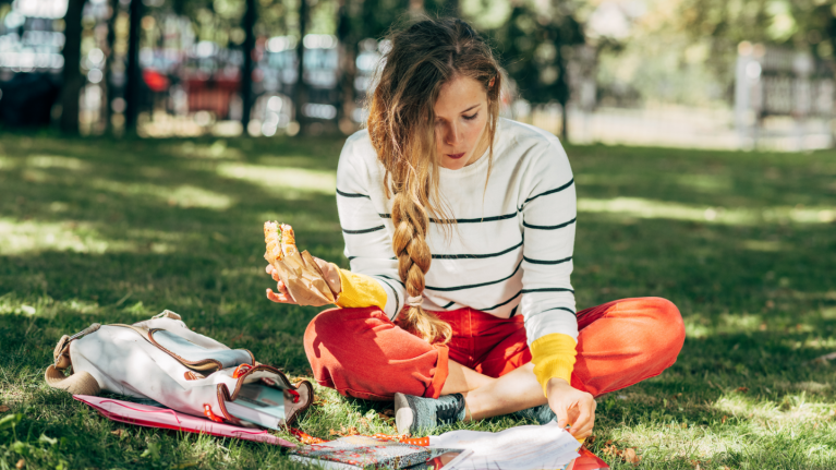 woman sitting on grass eating sandwich while reading